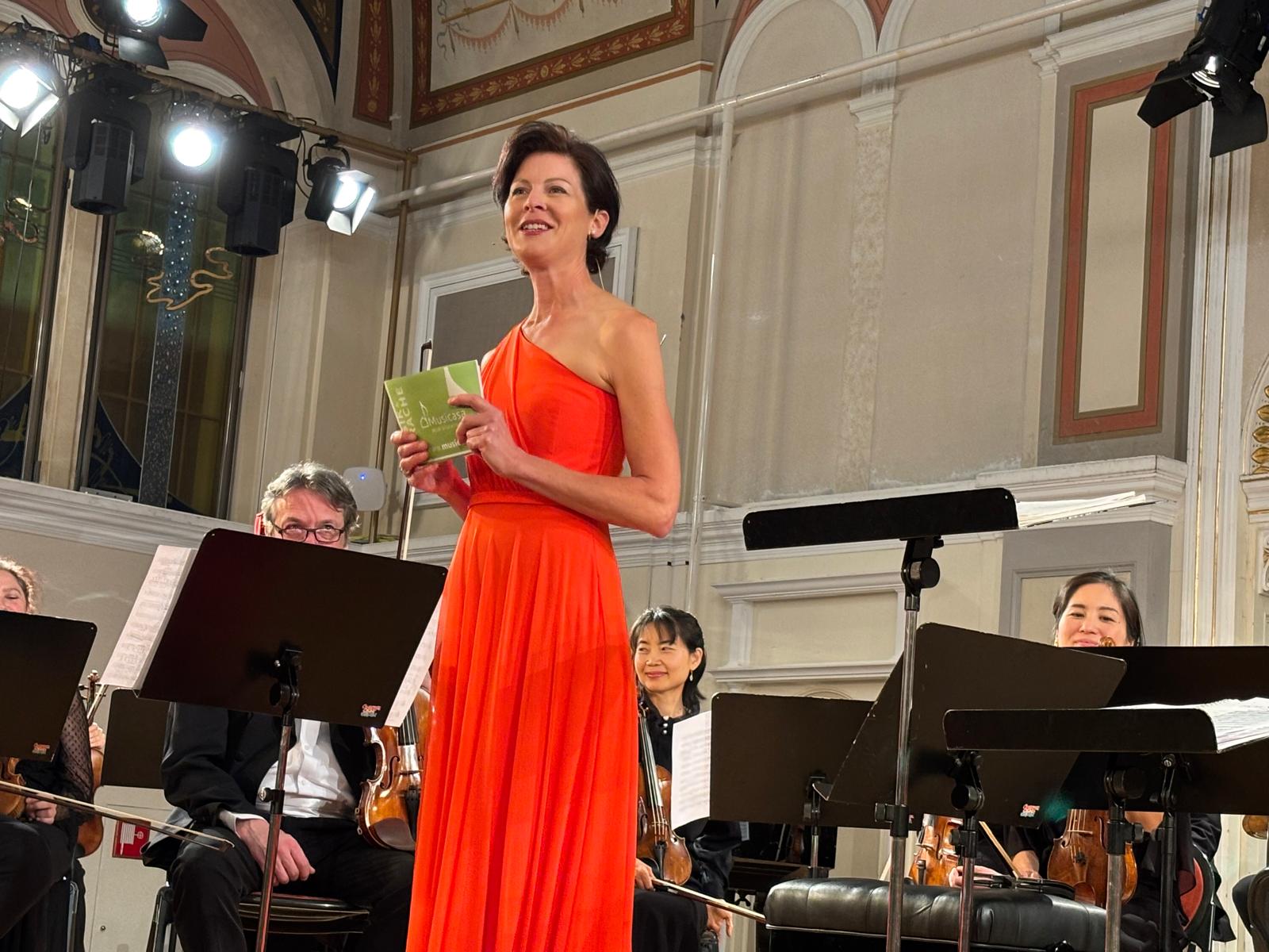 Frau (Katharina Göschke) in elegantem, orangefarbenem Kleid steht auf einer Bühne in einem prunkvollen Konzertsaal und spricht zum Publikum. Hinter ihr sitzen mehrere Musiker mit Streichinstrumenten vor Notenständern, der Raum ist mit Bögen und dekorativen Elementen gestaltet.